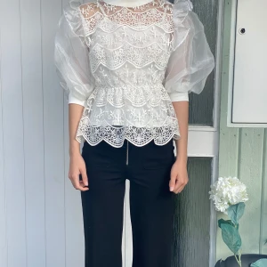Vit blus med spetsdetaljer - Elegant vit blus med spetsdetaljer och puffiga trekvartsärmar i transparent material. Blusen har en vacker broderad design och en feminin siluett med volangdetaljer i midjan.