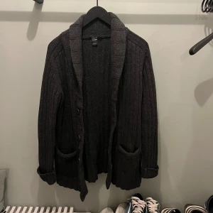 Mörkgrå stickad kofta från Flippa K - Säljer en mysig mörkgrå stickad kofta med knappar och stora fickor framtill. Perfekt för kyliga dagar och ger en avslappnad look. Koftan har långa ärmar och en ribbad krage.