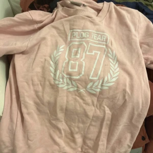 Rosa t-shirt med tryck - Säljer en rosa t-shirt med trycket 'Plog Year 87' och en lagerkrans. Perfekt för en avslappnad stil.