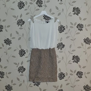 Vit och beige klänning med spetsdetaljer och glitter, Strl: S/36 - Elegant klänning med vit överdel och beige kjol. Kjolen har vackra spetsdetaljer och glittrande paljetter. Perfekt för festliga tillfällen med sin ärmlösa design och stilrena look.