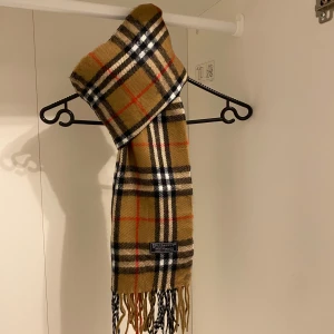 Burberry - Snygg och klassisk rutig halsduk från Burberry i beige. Den är Tillverkad i mjukt lammull  med fransar i ändarna. Perfekt för att hålla dig varm och stilren under kyliga dagar.