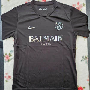 Svart PSG fotbollströja från Nike - Snygg svart PSG fotbollströja från Nike med Balmain Paris-logga på bröstet. Tröjan är kortärmad och har en rund halsringning. Perfekt för fotbollsfans som vill ha stil.