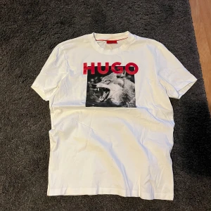 Hugo boss T-shirt  - Snygg vit t-shirt från Hugo med ett coolt vargmotiv och röd text på bröstet. Perfekt för en avslappnad stil med en touch av attityd. Kortärmad och i mjukt material.