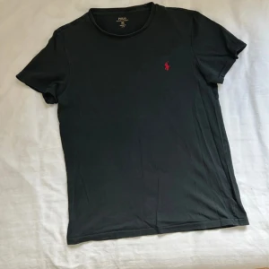 Svart t-shirt från Polo Ralph Lauren - Snygg svart t-shirt från Polo Ralph Lauren med röd broderad logga på bröstet. Perfekt för en stilren och avslappnad look. Tillverkad i mjukt material för extra komfort. Bara använd ett flertal gånger, bara att skriva vid frågor.