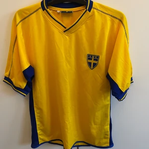 Gul fotbollströja från Heja Sverige - Säljer en gul fotbollströja från Heja Sverige med blå detaljer och korta ärmar. Tröjan har en V-ringad krage och ett broderat emblem på bröstet. Perfekt för att heja på Sverige i matcherna! 🇸🇪