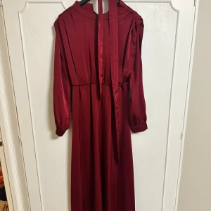Vinröd klänning med knytband - Elegant vinröd festklänning med långa ärmar och knytband vid midjan. Klänningen är använd några gånger men är i väldigt bra skick och bra kvalitet. 
