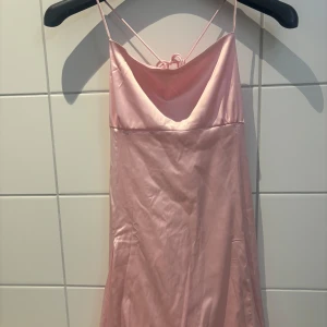 Rosa sidenklänning från Nelly - Elegant rosa klänning i siden från Nelly med smala axelband och v-ringad design. Klänningen har en öppen rygg med knytband för en justerbar passform. Perfekt för speciella tillfällen. Oanvänd med prislapp på.