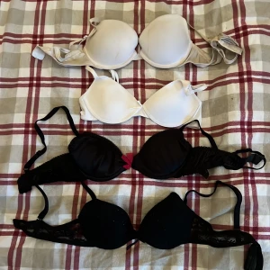 Fyra olika BH:ar i vitt och svart - Säljer fyra BH:ar i olika stilar och färger. Två vita och två svarta, varav en svart har en röd rosett och spetsdetaljer. Perfekta för olika tillfällen och outfits. Den längst ner svart push up storlek 70A, svart med rosett på 75A pusch up, vit med rosett 70AA, sista vit uppe 75a. 50kr/st 