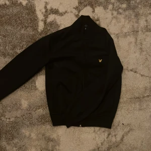  Lyle & Scott tröja - Snygg svart tröja från Lyle & Scott med en liten gul logga på bröstet. Tröjan har långa ärmar och en dragkedja framtill. Perfekt för en stilren look. Den är väldigt skön och snygg, använd fåtal gånger 