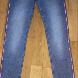 Blå jeans med färgglada sidor Tommy Hilfiger - Snygga Tommy Hilfiger blå jeans med en unik detalj av färgglada ränder längs sidorna. Perfekt för att ge din outfit en färgklick. Klassisk femficksdesign med knapp och dragkedja framtill.stl 164