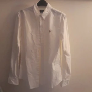 Vit skjorta från Ralph Lauren - Säljer en klassisk vit skjorta från Ralph Lauren med broderad logotyp på bröstet. Skjortan har långa ärmar och knappar framtill. Perfekt för en stilren look.