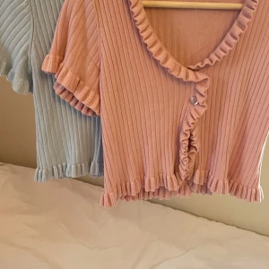 Topp med volangdetaljer - Säljer en söt ribbad topp med volangdetaljer i både rosa och ljusblått. Toppen har korta ärmar och knappar framtill. Var populära för några år sedan. 1 topp: 129kr. 2 toppar: 199kr ❤️Pris kan diskuteras❤️