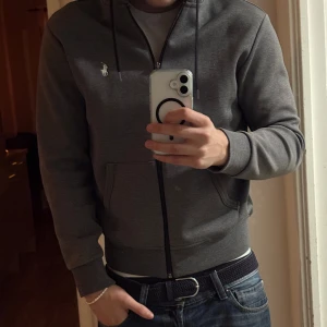 Grå hoodie från Polo Ralph Lauren - Säljer en stilren grå hoodie från Polo Ralph Lauren med dragkedja och broderad logga på bröstet. Perfekt för en avslappnad look. Den har långa ärmar och en bekväm passform. Den är köpt i butik. Den är i väldigt bra skick.