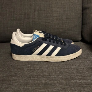 Adidas Gazelle i mörkblå mocka - Snygga Adidas Gazelle sneakers i mörkblå mocka med vita detaljer och klassiska tre ränder. Skorna har en vit sula och snörning framtill. Perfekta för en stilren och sportig look. Skick 9/10 lite smutsiga bara som enkelt borde gå bort skriv vid frågor OBS PRIS KAN DISKUTERAS