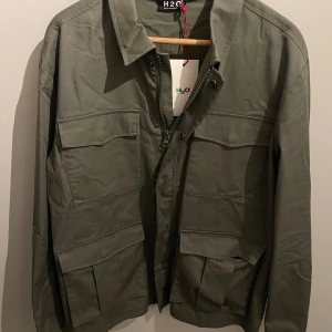 Grön jacka från H2O - Snygg grön jacka från H2O med dragkedja och flera praktiska fickor. Perfekt för en stilren look. Tillverkad i en bomulls- och nylonblandning för hållbarhet och komfort. Aldrig använd, fick den i julklapp men för stor för mig. Nypris 1200kr på booztlet!