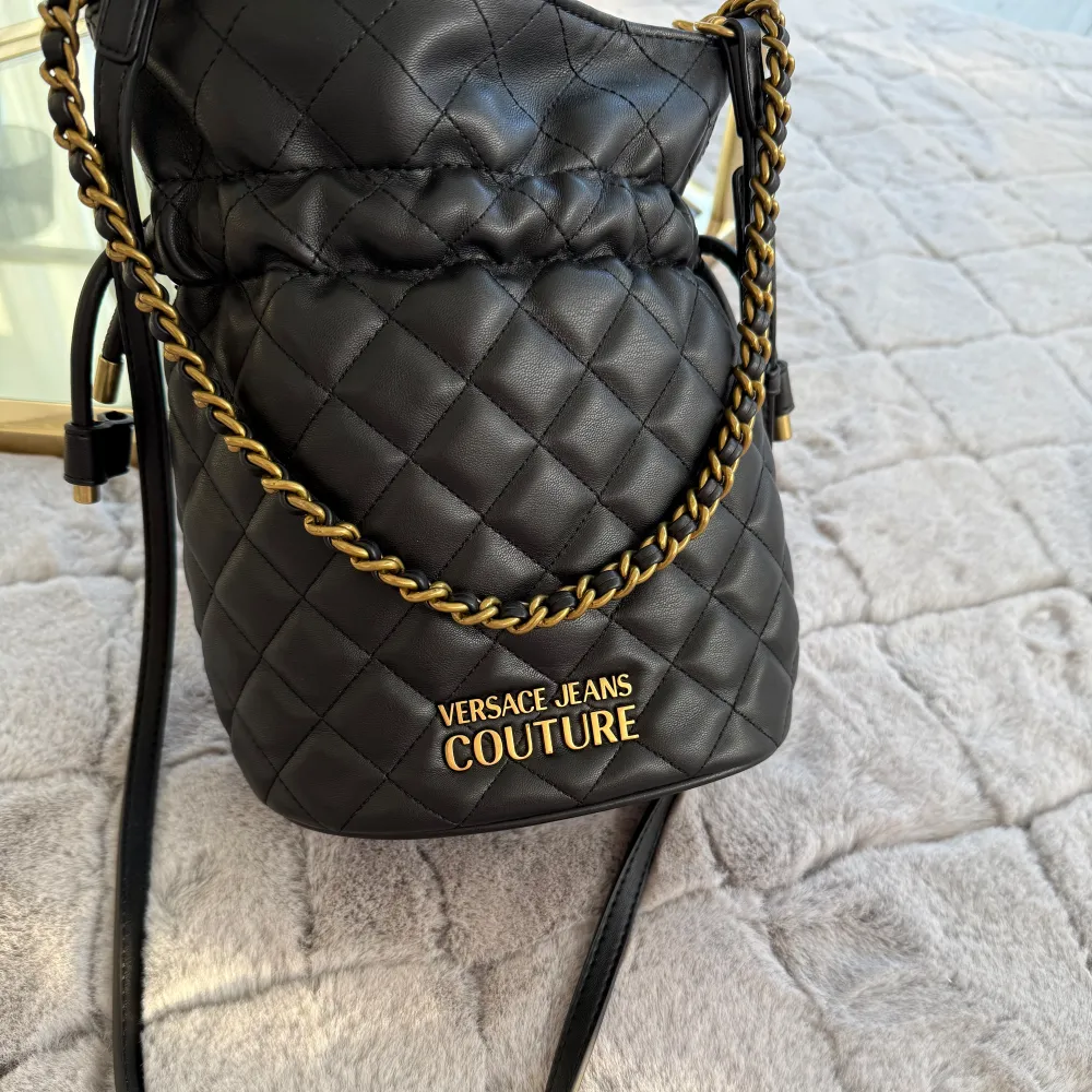 Snygg svart quiltad väska från Versace Jeans Couture med gulddetaljer. Väskan har en justerbar axelrem och en elegant kedja. Perfekt för att ge din outfit en lyxig touch.. Laukut & Käsilaukut.