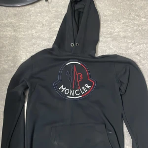 Svart hoodie från Moncler - Snygg svart hoodie från Moncler med stor broderad logga på framsidan. Tröjan har en klassisk passform med långa ärmar och en praktisk magficka. Perfekt för en stilren och bekväm look.