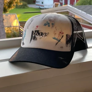 Svart och vit keps med konstnärligt mönster - Säljer en unik keps med en cool konstnärlig design. Kepsen är svart och vit med färgstänk i olika nyanser och texten 'NART' broderad på framsidan. Den har en nätbaksida för bra ventilation och en justerbar rem för perfekt passform. Perfekt för sommarens alla äventyr!