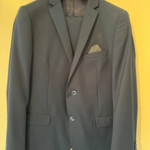 Mörkblå kostym från Between - Säljer en elegant mörkblå kostym från Between. Kavajen har en klassisk design med två knappar och en stilren bröstficka med en mönstrad näsduk. Byxorna matchar perfekt och har en rak passform. Perfekt för både formella tillfällen och fest.