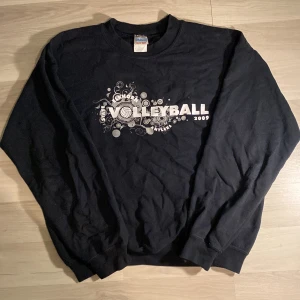 Svart sweatshirt från Gildan - Säljer en svart sweatshirt från Gildan med tryck på både fram- och baksidan. På framsidan står det 'Volleyball 2009' med ett coolt grafiskt mönster. Baksidan listar namn och nummer, vilket ger en unik och personlig touch. Tröjan är i en bekväm och varm 'Heavy Blend' material, perfekt för kyliga dagar. Passar bra för både vardag och sportiga aktiviteter.
