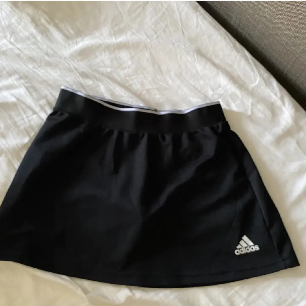 Säljer en snygg och bekväm svart sportkjol från Adidas. Perfekt för träning eller tennis! Kjolen har en elastisk midja för bästa passform och är gjord i ett lätt och andningsbart material. Adidas-loggan syns diskret i hörnet. Passar perfekt för en aktiv livsstil!. Kjolar.