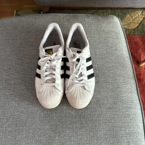 Vita Adidas Superstar sneakers - Säljer ett par vita Adidas Superstar sneakers i storlek 43. Skorna är i bra skick med några tecken på användning. De har den klassiska svarta tre-randiga designen och snörning framtill. Perfekta för både vardag och träning! Priset är ej hugget i sten
