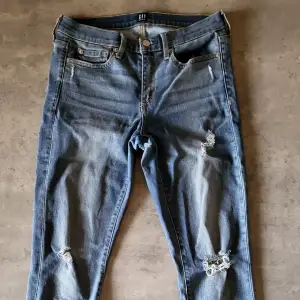 Säljer ett par blå jeans från GAP i storlek M. De är i en klassisk blå färg med slitningar på framsidan och baksidan, vilket ger dem en cool och avslappnad look. Jeansen har en knapp och dragkedja framtill samt fem fickor. Perfekta för en casual outfit!