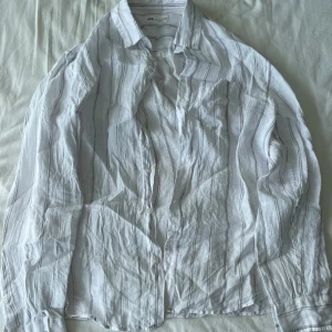 Vit och grå randig skjorta från Zara - Säljer en stilren vit skjorta med grå ränder från Zara. Skjortan har långa ärmar och knappar framtill. Har använts sparsamt och är i 100% linne. Perfekt för både vardag och fest! 🕺Frågor? Skriv!