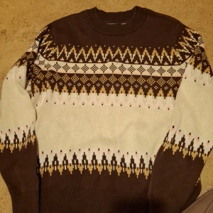 Brun och vit stickad tröja med mönster - Säljer en mysig brun och vit stickad tröja med ett snyggt geometriskt mönster. Perfekt för kalla dagar! Tröjan har långa ärmar och ribbade muddar. Det finns en liten fläck på framsidan, men annars i bra skick. Perfekt för höst och vinter!