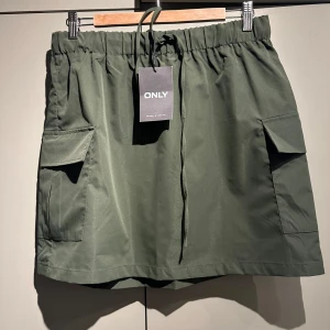 Grön cargokjol från ONLY - Säljer en snygg grön cargokjol från ONLY i storlek L. Kjolen har praktiska fickor på sidorna och en bekväm resårmidja med dragsko. Perfekt för en avslappnad och trendig look. Passar bra till både vardag och utflykter. 🌿