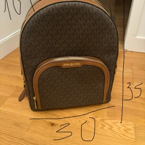 Brun ryggsäck från Michael Kors - Säljer en helt ny, snygg brun ryggsäck från Michael Kors i mycket bra skick. Väskan har ett klassiskt MK-mönster och detaljer i brunt skinn. Den har en praktisk dragkedja och en framficka för extra förvaring. Perfekt för både vardag och resa!
