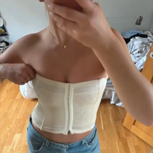 Vit korsett med blommigt mönster - Säljer en elegant vit korsett med ett subtilt blommigt mönster. Den är axelbandslös och har en tight passform som framhäver figuren. Perfekt för festliga tillfällen. Bör passa både S och M. Har en fläck på ryggen som kanske kan tas bort