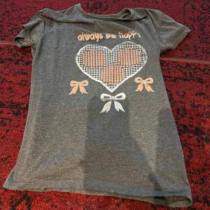 Säljer en söt grå t-shirt med texten 'always be happy' och ett stort hjärta i mitten. T-shirten har korta ärmar och är dekorerad med små rosetter i orange och vitt. Perfekt för en avslappnad dag eller sommarhäng!