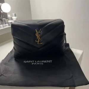 Väska från Saint Laurent modellen Toy - Säljer en elegant svart quiltad väska från Saint Laurent. Väskan har en lyxig känsla med det ikoniska YSL-emblemet i guld på framsidan. Den är perfekt för både vardag och fest. Insidan har flera fack och en dragkedja för säker förvaring. Kommer med originalpåse.
