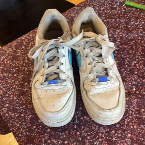 Nike Air Force i vitt och blått - Säljer ett par klassiska Nike Air Force sneakers i vitt med blå detaljer. Skorna har snörning och är i acceptabelt skick med lite slitage. Perfekta för vardagsbruk och ger en sportig look. Storlek är 38.5 perfekt passform.