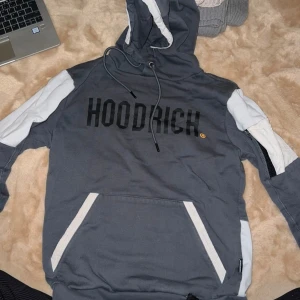 Grå hoodie från Hoodrich - Säljer en grå hoodie från Hoodrich i mycket bra skick. Den har en cool design med svart text på framsidan och en världskarta i guld på baksidan. Hoodien har en stor ficka fram och vita detaljer på ärmarna. Perfekt för höst och vinter!