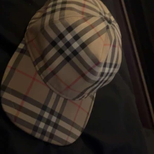 Burberry keps  - Snygg rutig keps från Burberry i klassiskt mönster med beige, svart och röda rutor. Perfekt för att ge din outfit en stilren touch. Kepsen är i bra skick och passar för både vardag och fest. Justerbar passform för extra komfort.