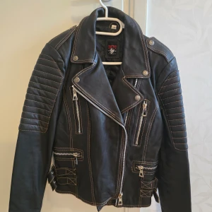 Svart skinnjacka med dragkedjor - Säljer en snygg svart skinnjacka från Urban Republic i storlek M. Jackan har coola dragkedjedetaljer och quiltade axlar. Perfekt för höst och vår, med en edgy stil som passar både till vardags och fest. Den är i mycket bra skick och redo för nya äventyr!