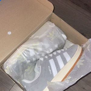 Grå Adidas Campus sneakers - Säljer ett par snygga grå Adidas Campus sneakers i nytt skick. De har den klassiska tre-randiga designen i vitt och en stilren gummisula. Perfekta för både vardag och fest! Skorna kommer i originalkartong med Adidas-logga på pappret.Köpte skorna för 1700kr använda 1gång kvitto borde finnas 