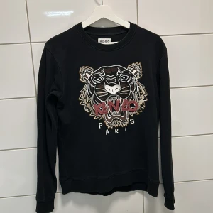 Svart tröja med tigerbroderi från Kenzo - Säljer en svart tröja från Kenzo med ett coolt tigerbroderi på framsidan. Tröjan har långa ärmar och är i mycket bra skick. Perfekt för både vardag och fest! 🐯