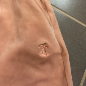 Rosa mjukisbyxor från Juicy Couture - Säljer ett par superbekväma rosa mjukisbyxor från Juicy Couture i storlek S. De har en elastisk midja med snörning och ett broderat emblem på sidan. Perfekta för en mysig dag hemma eller en avslappnad utflykt. 🩷