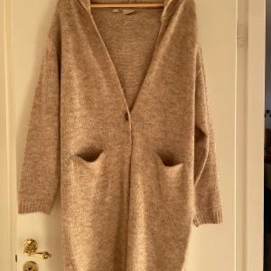 Beige stickad kofta från Bodyflirt - Mysig beige stickad kofta från Bodyflirt i mycket bra skick. Den har en luva och två stora fickor framtill, perfekt för kyliga höst- och vinterdagar. Koftan är långärmad och har en avslappnad passform. Perfekt att slänga på sig för en casual look!