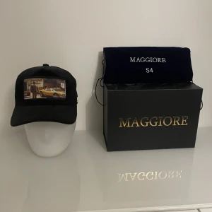Svart keps från Maggiore - Snygg svart keps från Maggiore med ett coolt tryck av en bil på framsidan. Kepsen har justerbar rem baktill för perfekt passform. Perfekt för bilentusiaster eller som en stilren accessoar till vardagsoutfiten.