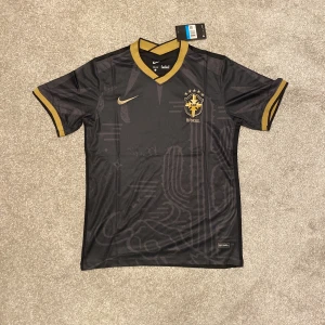 Svart Brasilien fotbollströja från Nike - Säljer en snygg svart Brasilien fotbollströja från Nike i Dri-FIT material. Tröjan har korta ärmar och guldiga detaljer vid krage och ärmslut. Perfekt för fotbollsfans som vill ha något unikt! 🖤