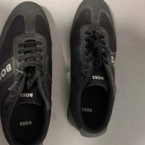 Snygga svarta sneakers från Boss i mycket bra skick. De har en stilren design med snörning och Boss-loggan tydligt på sidan. Perfekta för både vardag och fest. Skorna är gjorda i en kombination av skinn och syntetmaterial för en hållbar och bekväm passform.