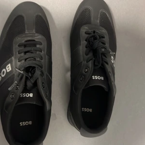 Svarta sneakers från Boss - Snygga svarta sneakers från Boss i mycket bra skick. De har en stilren design med snörning och Boss-loggan tydligt på sidan. Perfekta för både vardag och fest. Skorna är gjorda i en kombination av skinn och syntetmaterial för en hållbar och bekväm passform.