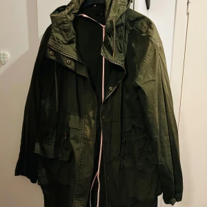 Grön parkajacka med huva - Säljer en grön parkajacka i bra skick. Jackan har en praktisk huva och flera fickor med dragkedja. Perfekt för höst och vår, med en skön passform som är normal. Den är långärmad och har en dragsko i midjan för justerbar passform.