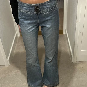 Jeans med läder snörning - Säljer ett par snygga blå jeans med bootcut-stil och mid-rise. De har en cool snörning framtill och klassiska bakfickor med knappdetaljer. Midjemåttet rakt över är 35cm och innerbenslängden är 73cm.