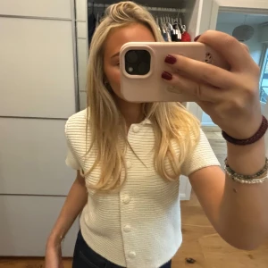 Vit stickad kortärmad topp - Säljer en superfin vit ribbad topp med korta ärmar. Den har knappar framtill och en klassisk rund halsringning. Perfekt för både vardag och festliga tillfällen. Toppen är i mycket bra skick och passar perfekt till jeans eller kjol.