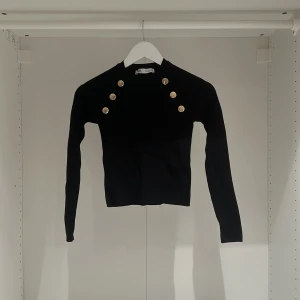 Svart ribbad tröja från Zara - Säljer en snygg svart ribbad tröja från Zara i storlek S. Den har långa ärmar och coola guldknappar på axlarna som ger en extra touch. Perfekt för både vardag och fest! 🖤